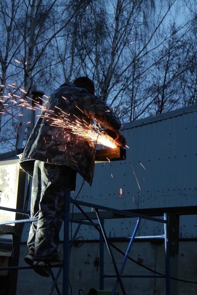 Arbeiter mit einer Schneidemaschine in den Händen auf einem Gerüst schneidet Metall, ein Bündel heller Funken zerstreut sich in der Dämmerung niedrigen Blickwinkels foto