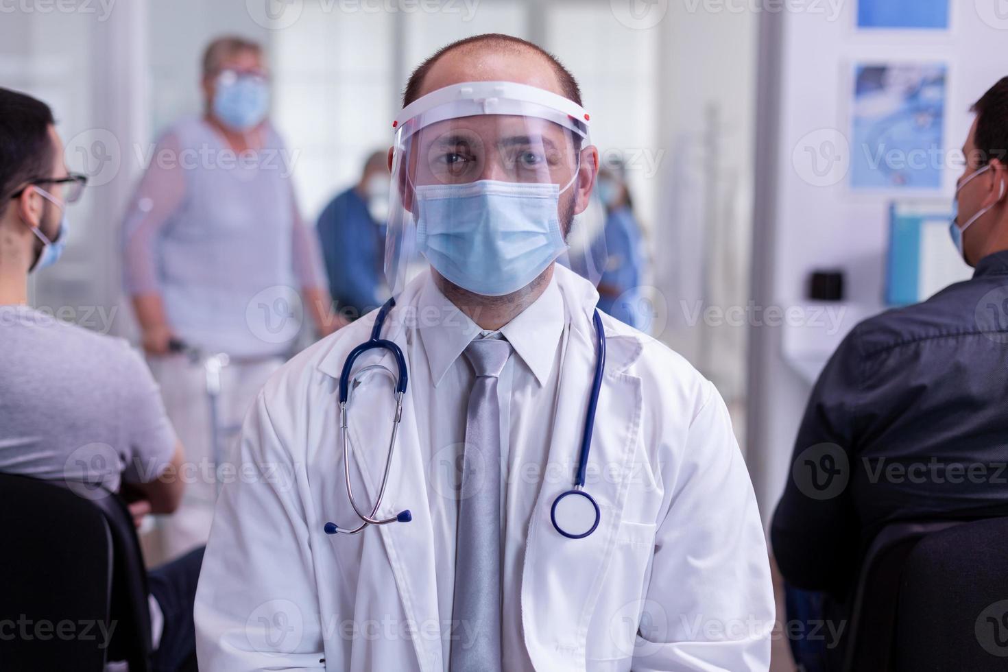 Arzt mit Maske, der im Empfangsbereich des Krankenhauses sitzt und in die Kamera schaut foto