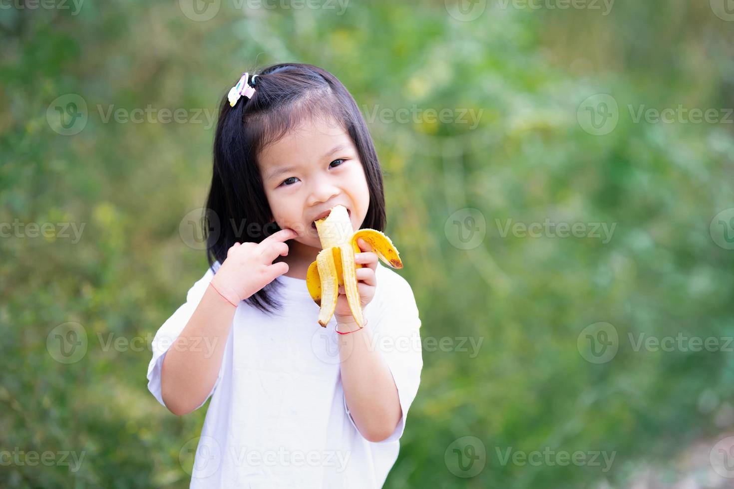 Portrait Kind Mädchen 4-5 Jahre alt süße gelbe Banane beißen. natürlicher grüner Hintergrund. foto