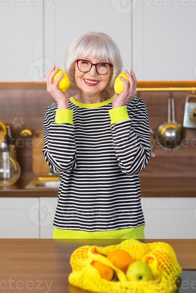 Fröhliche hübsche ältere lächelnde Frau in gestreiftem Pullover, die Zitronen für Limonade hält, während sie in der Küche steht. gesunder, saftiger Lebensstil, Zuhause, Seniorenkonzept. foto