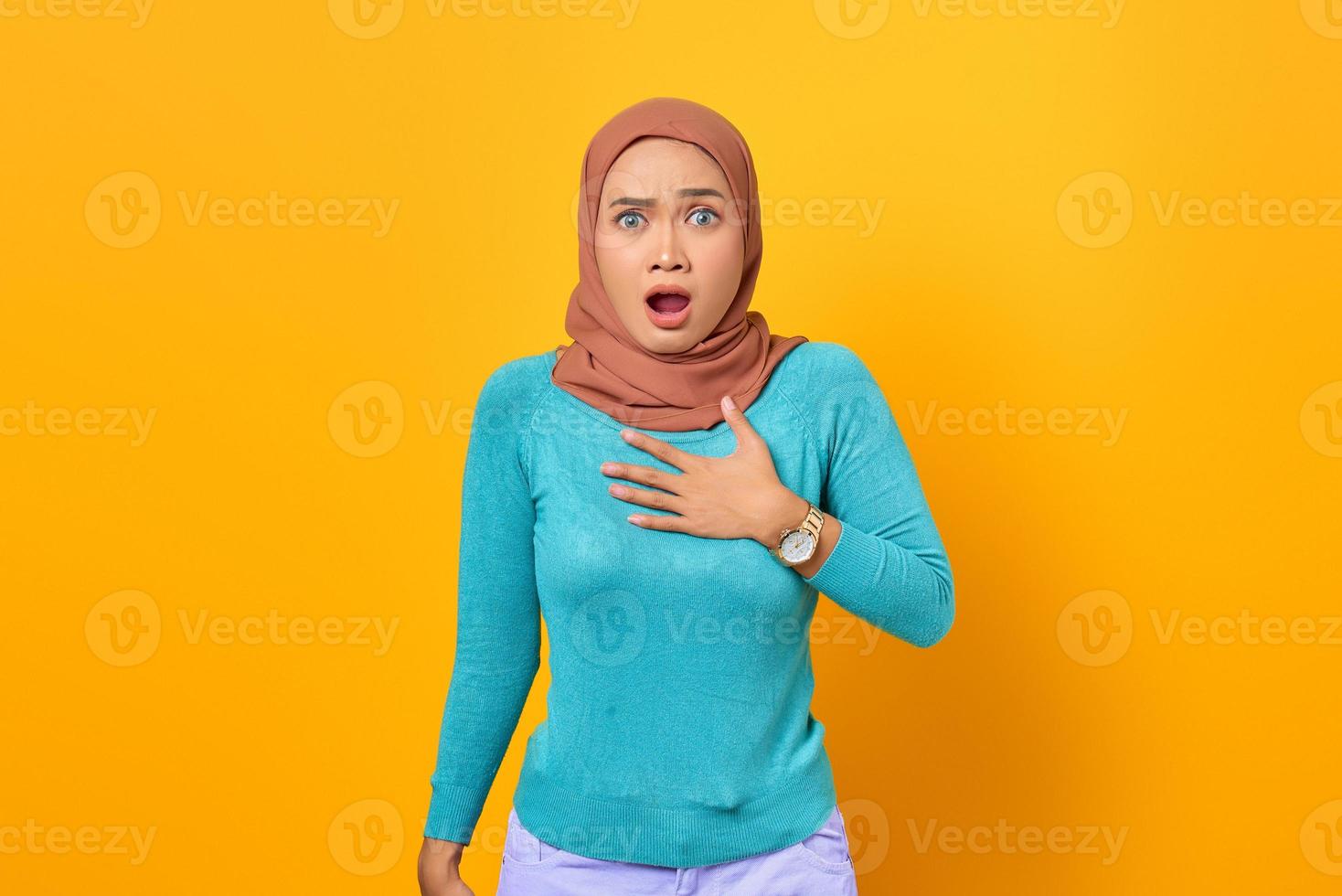 schockierte junge asiatische frau, die eine hand auf einer brust mit offenem mund auf gelbem hintergrund hält foto