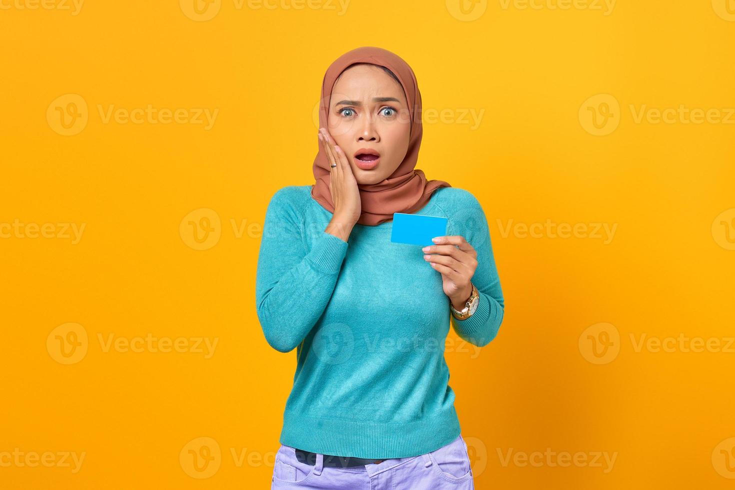 schockierte asiatische frau, die kreditkarte mit offenem mund und hand auf einer wange auf gelbem hintergrund hält foto