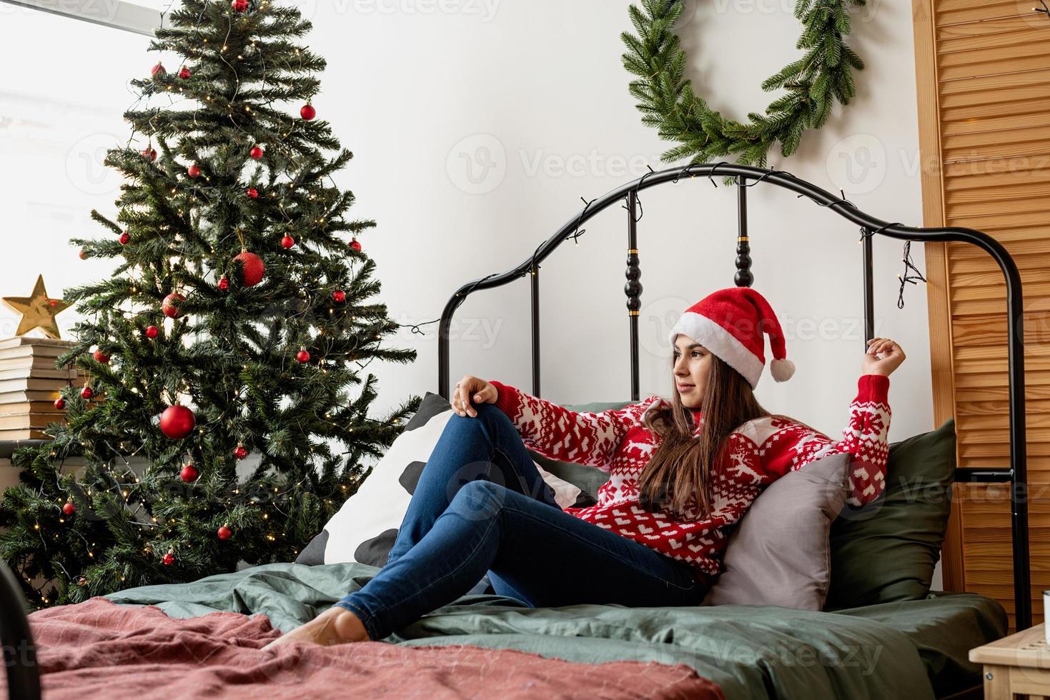 junge Frau in rotem Pullover und Weihnachtsmütze sitzt auf dem Bett und feiert Weihnachten foto