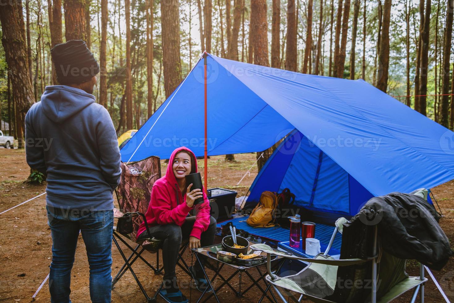 Junge asiatische Paare, die das Camping im Kiefernwald genießen, sitzen und essen auf der Campingseite inmitten der Natur. foto