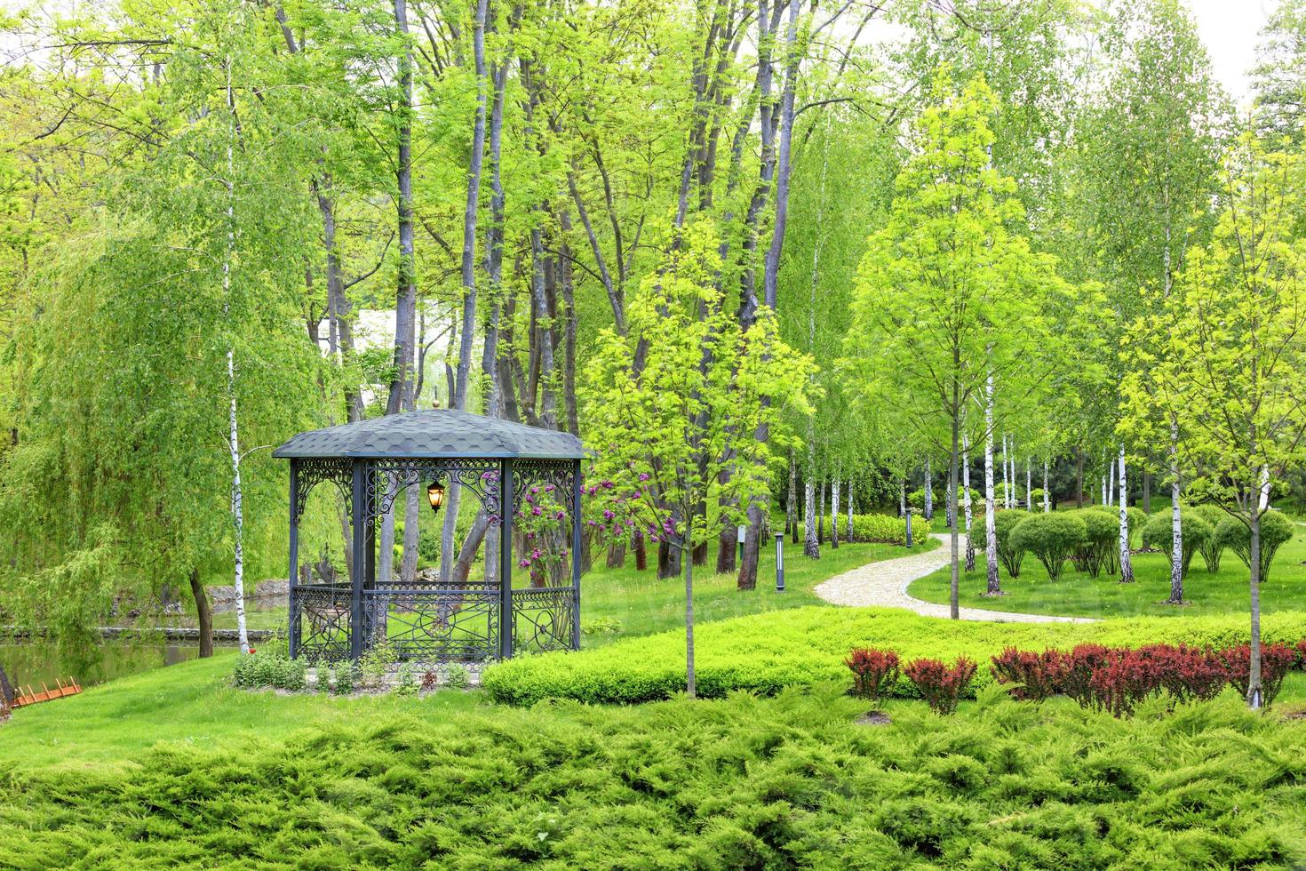 durchbrochene Metalllaube mit geschmiedeten Elementen, umgeben von einem wunderschönen Frühlingspark mit Landschaftsdesign. foto