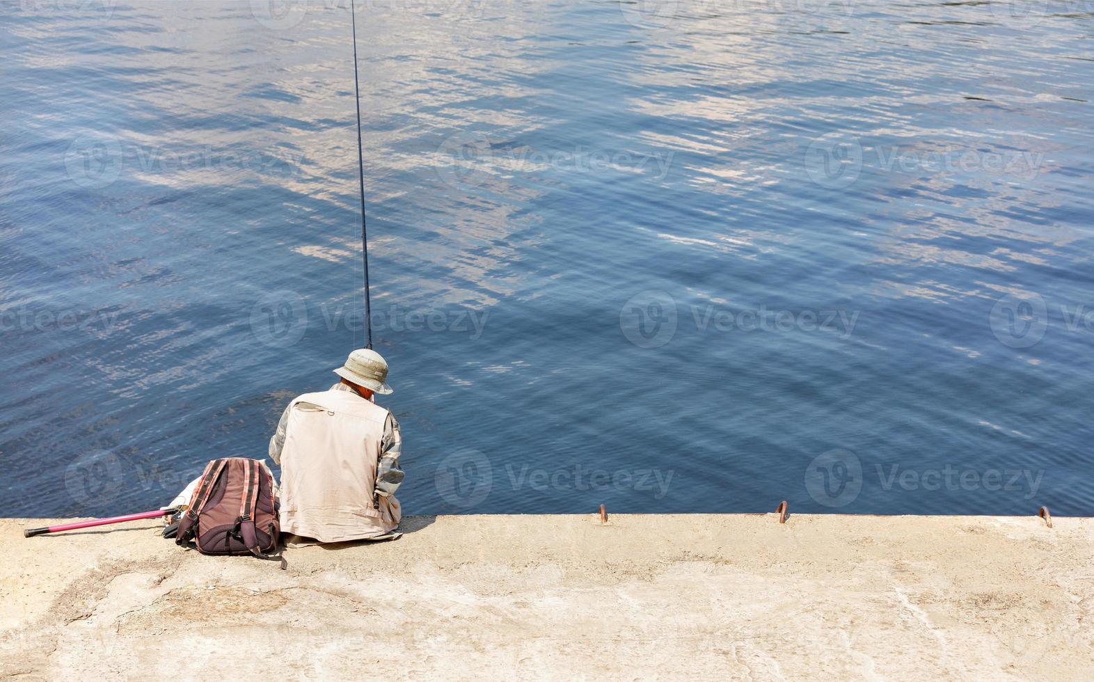 Ein einsamer Fischer sitzt an einem sonnigen Sommertag auf einem Betonpier mit einer Angelrute in der Nähe des Wassers. foto
