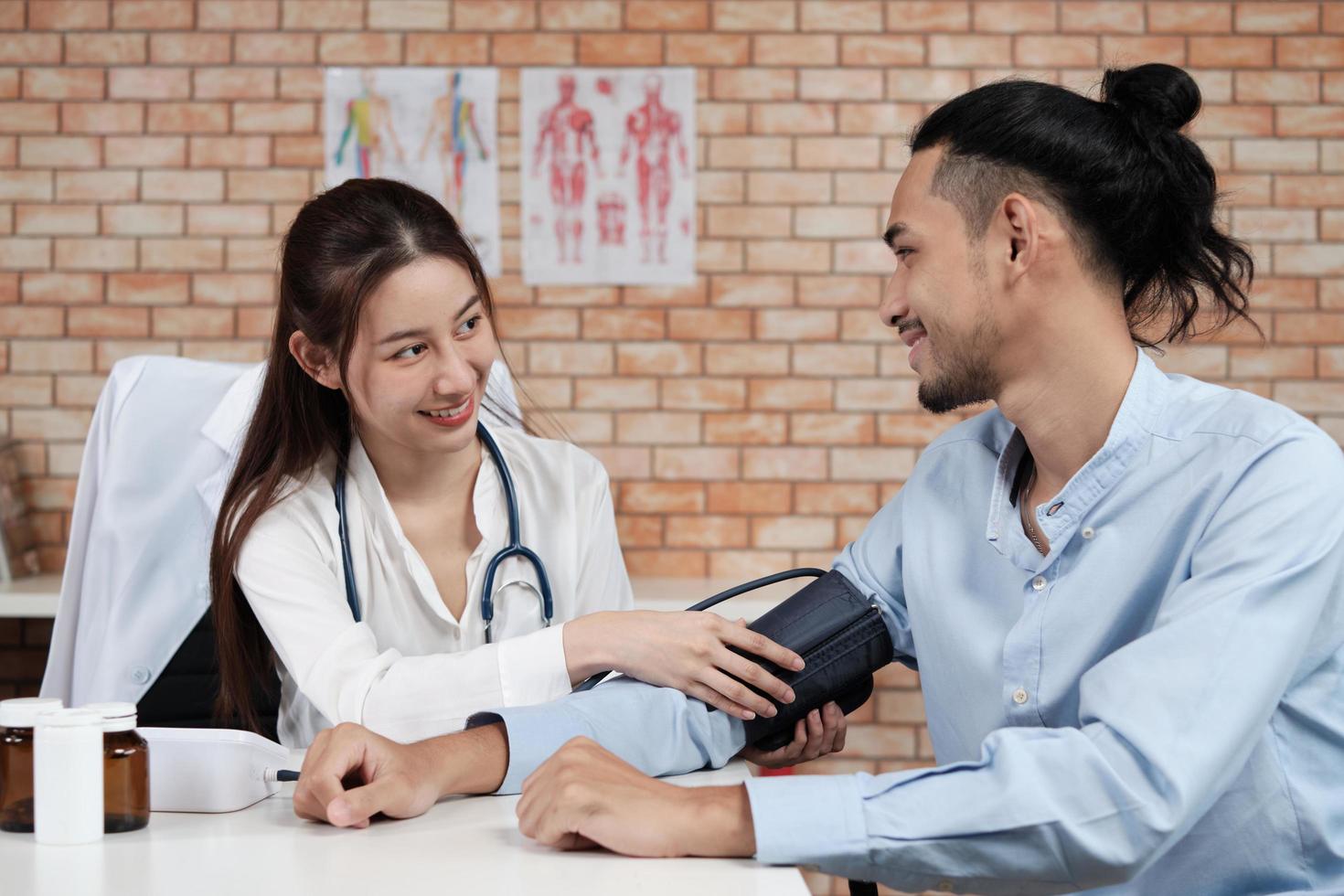 Schöne Ärztin in weißem Hemd, die eine asiatische Person mit Stethoskop ist, ist eine Gesundheitsprüfung eines männlichen Patienten in einer medizinischen Klinik mit Backsteinmauerhintergrund und lächelt, berät den Beruf des Facharztes. foto