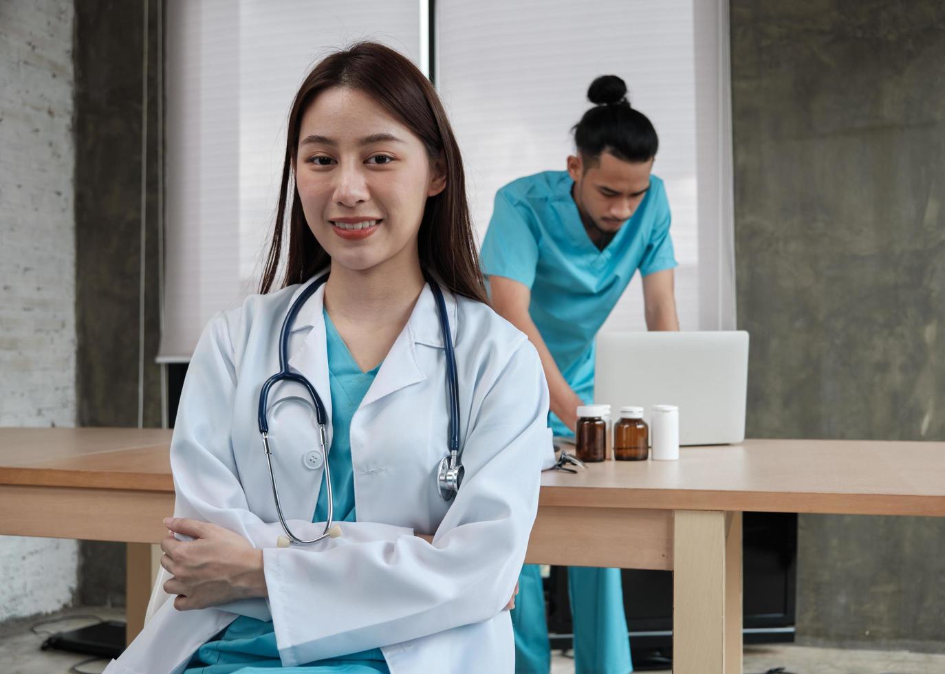 Porträt einer schönen Ärztin asiatischer Abstammung in Uniform mit Stethoskop. Lächeln und Blick in die Kamera in einer Krankenhausklinik, männlicher Partner, der hinter ihr arbeitet, zwei professionelle Personen. foto