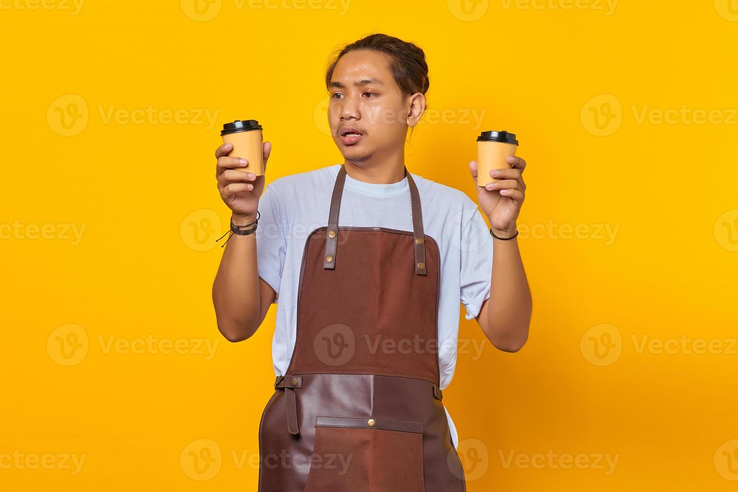 Porträt eines verwirrten gutaussehenden Mannes, der zwei Tassen Kaffee hält und nach vorne schaut, isoliert auf gelbem Hintergrund foto