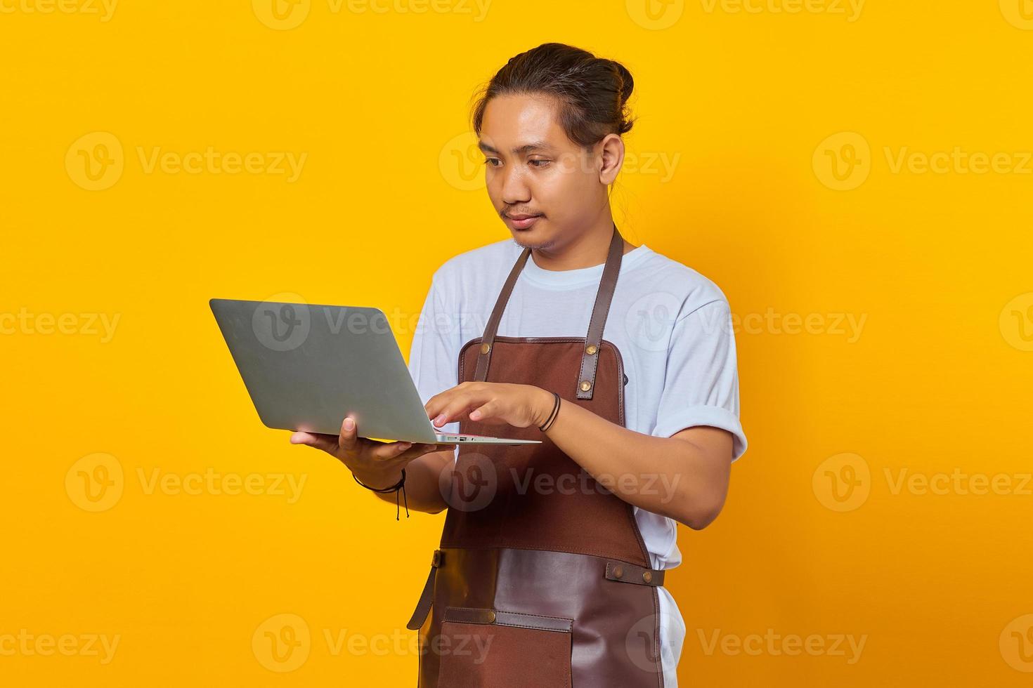 Porträt eines gutaussehenden jungen Barista mit Laptop, der ernsthaft Büroarbeit auf gelbem Hintergrund macht foto