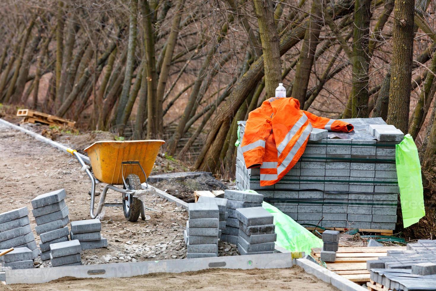 Die orangefarbene Jacke eines Baumeisters liegt auf einem Stapel von Pflastersteinen in der Nähe einer gelben Schubkarre vor dem Hintergrund wachsender Bäume im Park. foto