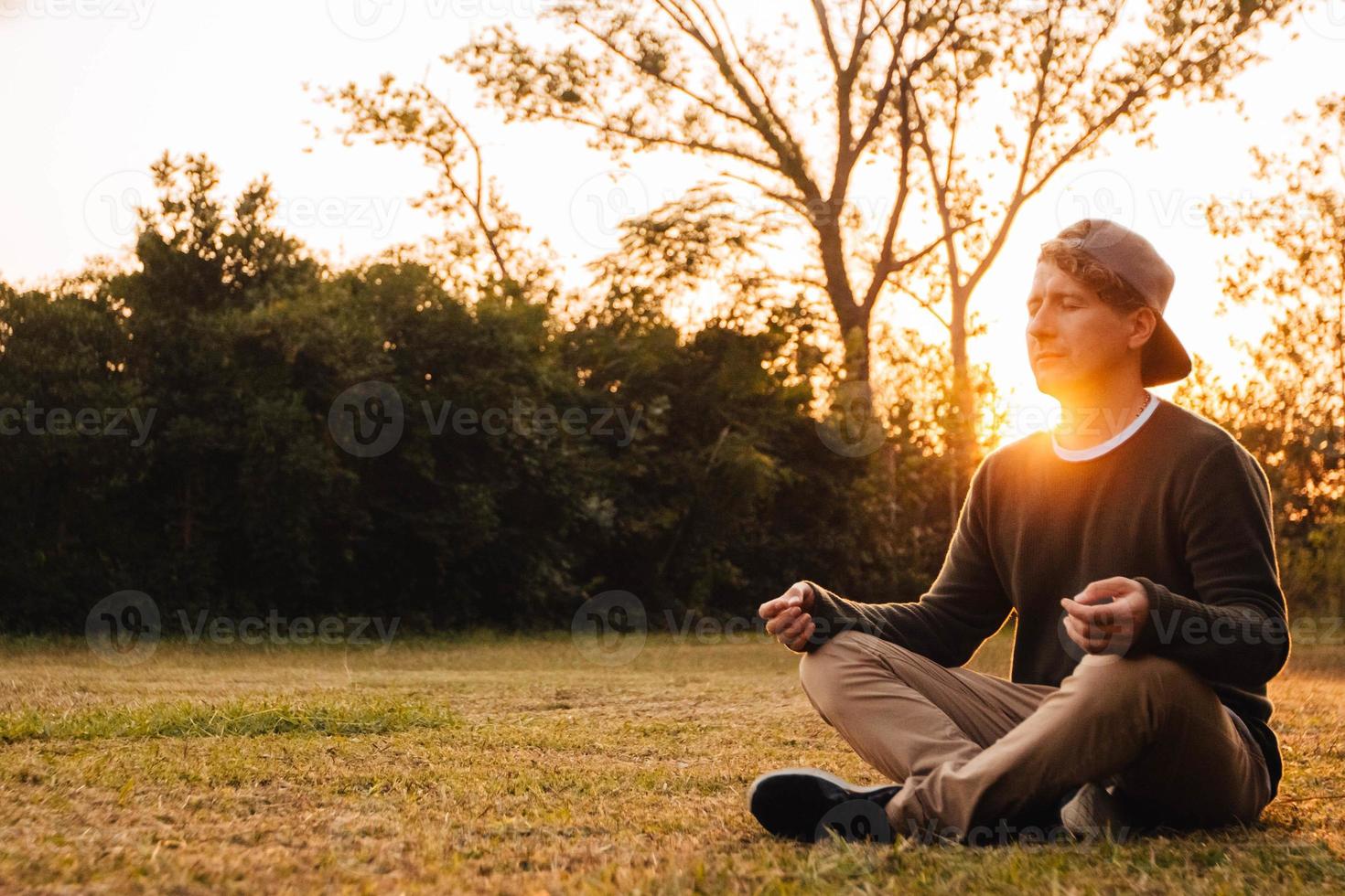 Mann in meditativer Position sitzt auf einer Wiese vor dem Hintergrund von Wald und Sonnenuntergang foto