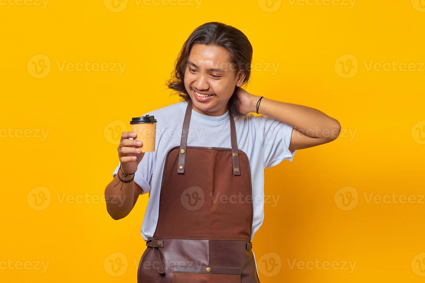 Porträt eines gutaussehenden Mannes mit Schürze, der auf eine Tasse Kaffee schaut, die isoliert auf gelbem Hintergrund gehalten wird foto
