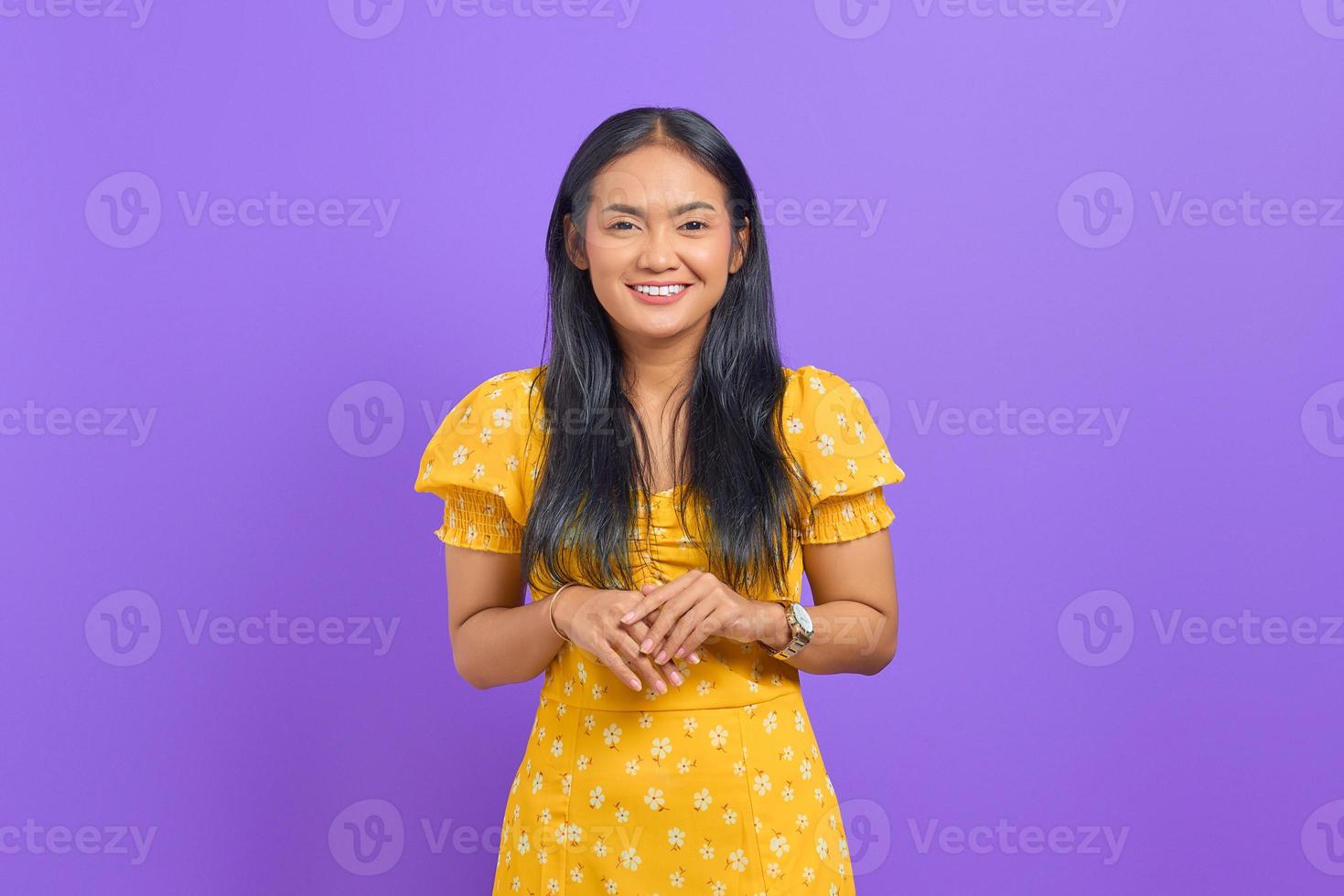 lächelnde junge asiatische frau hält die hand zusammen und fühlt sich optimistisch auf lila hintergrund foto