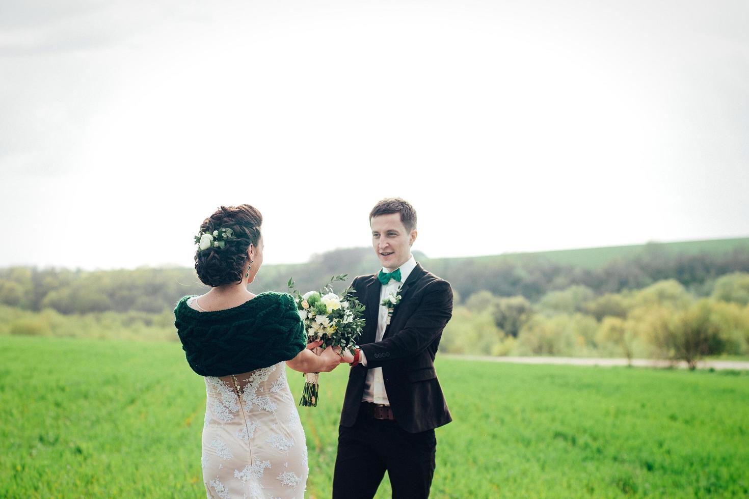 der Bräutigam im braunen Anzug und die Braut im elfenbeinfarbenen Kleid auf grüner Wiese foto