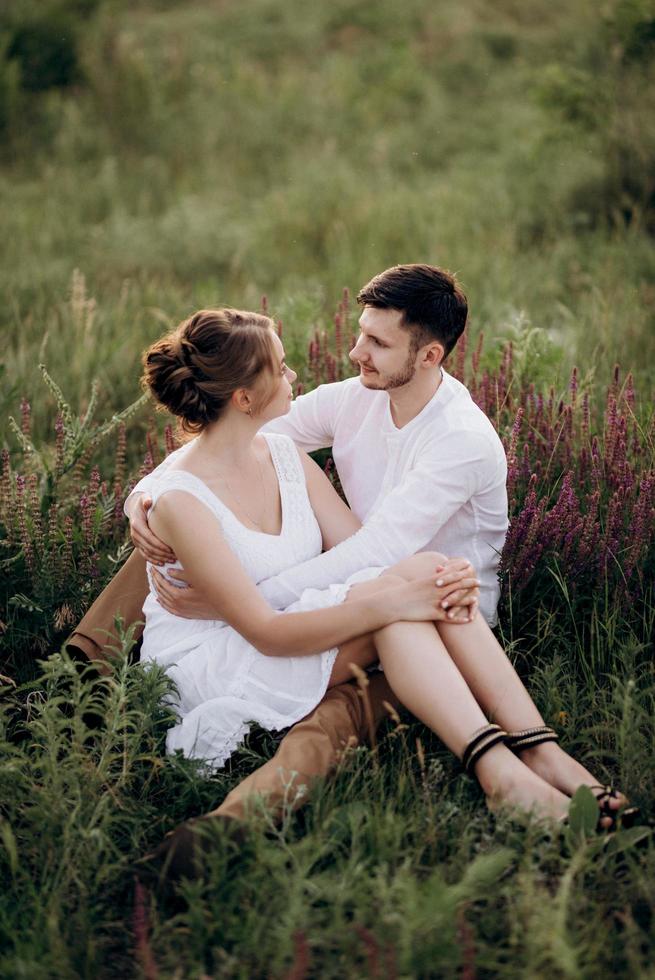 Mädchen in einem weißen Sommerkleid und ein Typ in einem weißen Hemd auf einem Spaziergang bei Sonnenuntergang mit einem Blumenstrauß foto