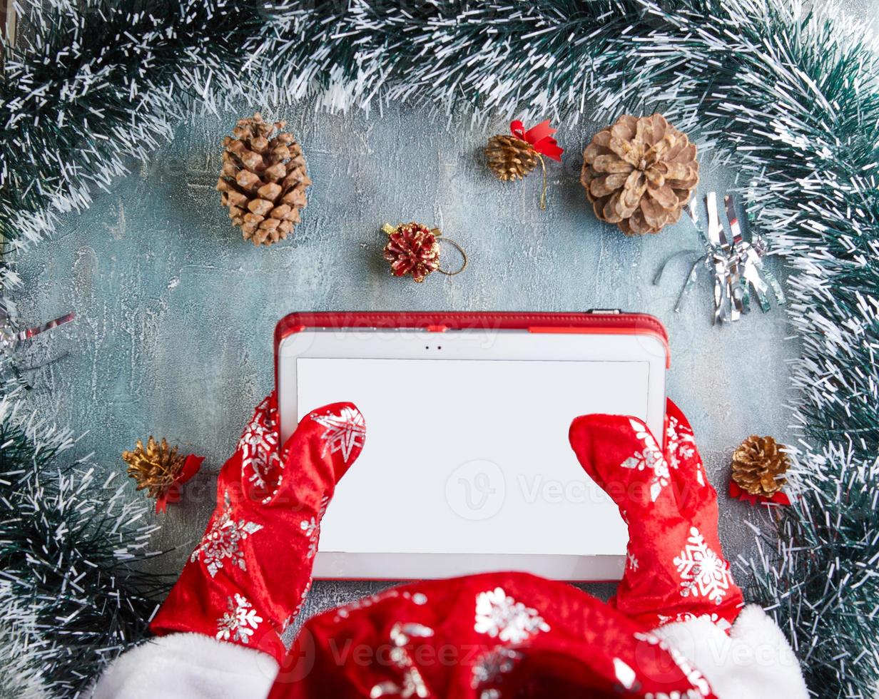 Frohe Weihnachten und ein glückliches neues Jahr-Konzept. Der Weihnachtsmann sitzt am Neujahrstisch mit einem Tablet in der Hand foto