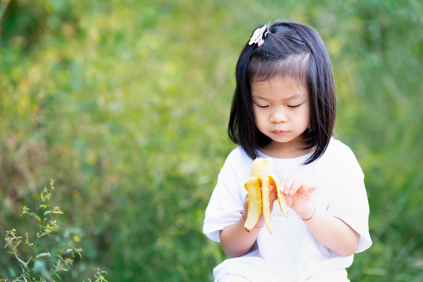 süßes Mädchen schält köstliche Banane, die sie in der Hand hält. grüner Naturhintergrund. foto