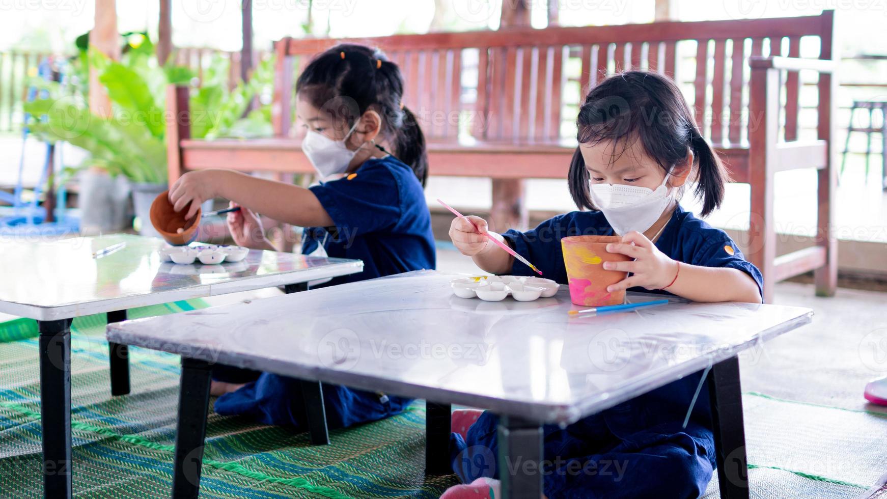 Kindergartenkind Mädchen lernt Kunstmalerei mit Aquarell und Pinsel auf kleinem Tisch, während sie an einer Exkursion teilnimmt. Kinder tragen weiße chirurgische Gesichtsmasken. foto