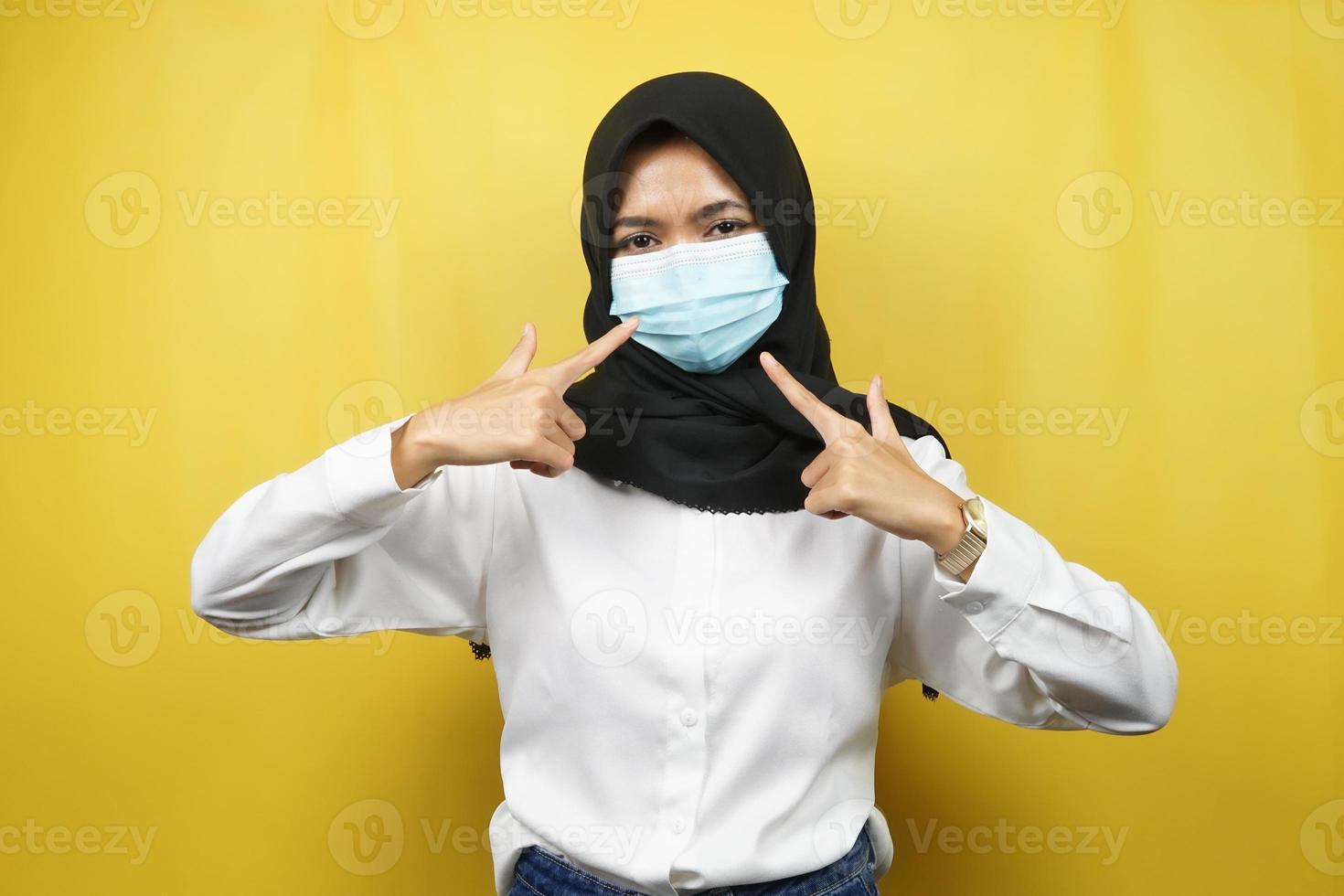 muslimische Frau mit medizinischer Maske mit Hand, die auf Maske zeigt, Anti-Corona-Virus-Bewegung, Anti-Covid-19-Bewegung, Gesundheitsbewegung mit Maske, einzeln auf gelbem Hintergrund foto