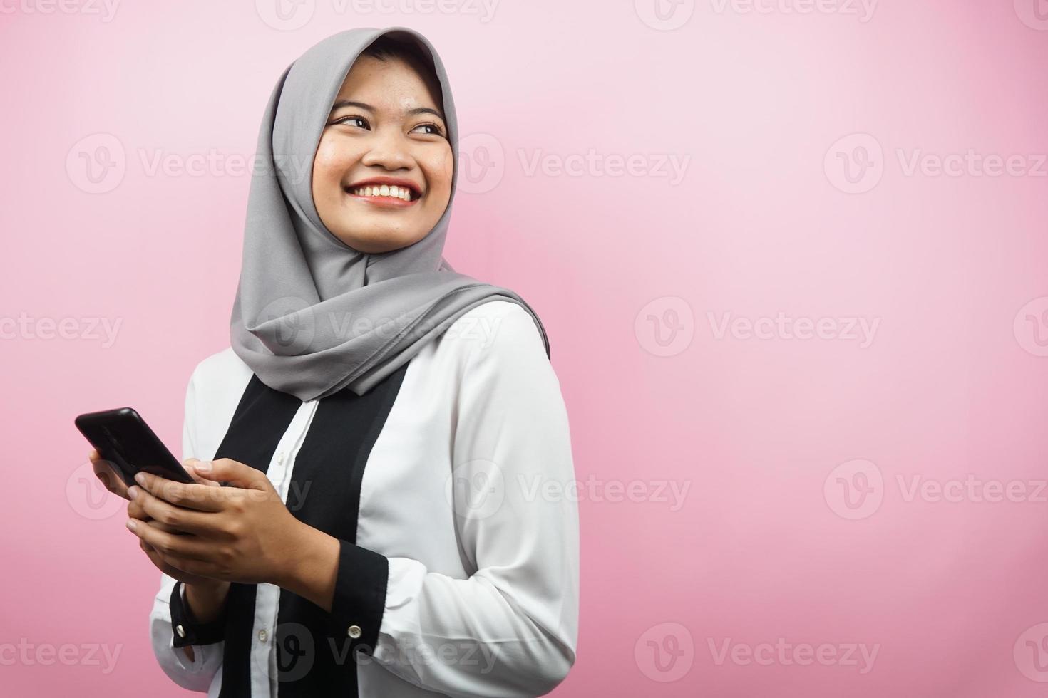 Schöne junge asiatische muslimische Frau, die selbstbewusst, enthusiastisch und fröhlich lächelt, mit den Händen, die Smartphone halten, Blick auf den leeren Raum, der etwas präsentiert, einzeln auf rosafarbenem Hintergrund foto