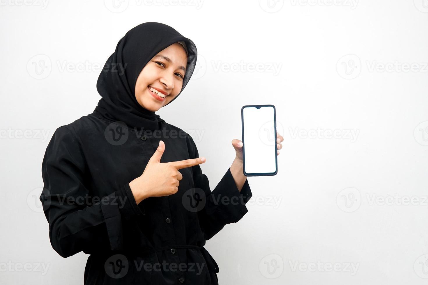 schöne junge asiatische muslimische frau mit hand, die auf smartphone zeigt, etwas fördert, hand, die app fördert, selbstbewusst, enthusiastisch und fröhlich lächelt, isoliert auf weißem hintergrund foto