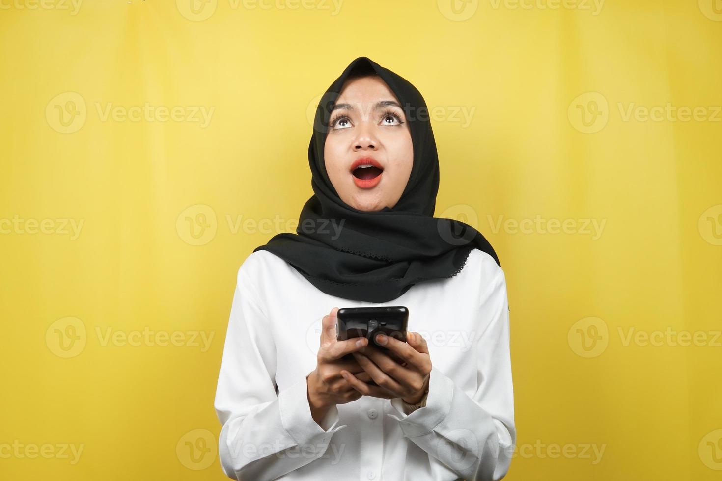 schöne junge asiatische muslimische frau schockiert, überrascht, wow-ausdruck, hand, die smartphone hält, nach oben schauend, isoliert auf gelbem hintergrund foto