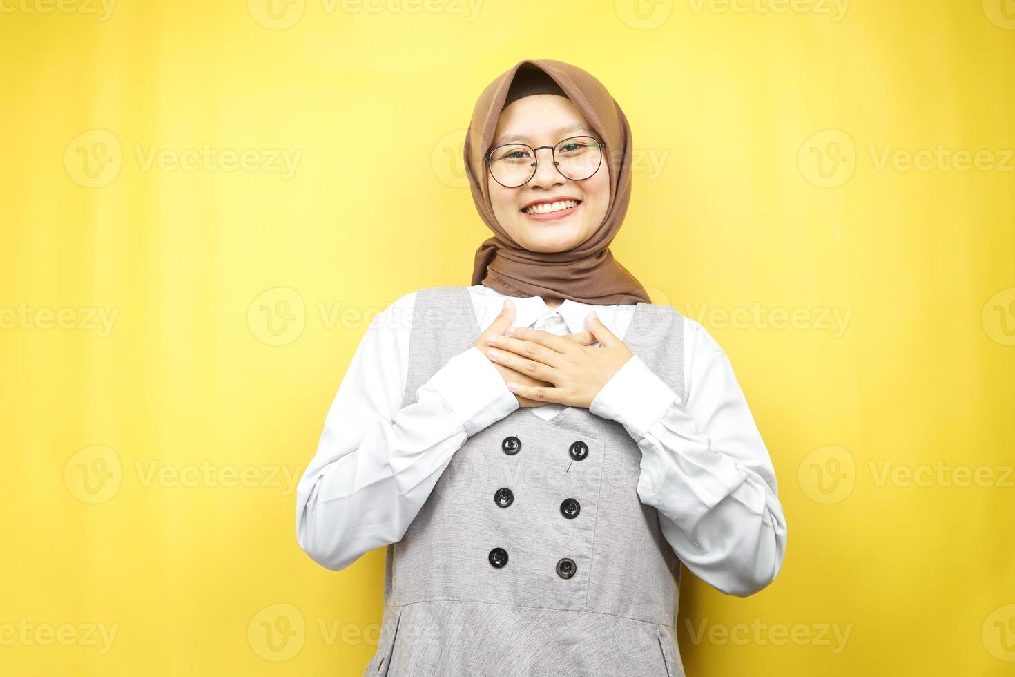 schöne junge asiatische muslimische frau, die überrascht und fröhlich lächelt, mit den händen, die die brust halten, aufgeregt, nicht erwartet, die kamera einzeln auf gelbem hintergrund betrachtend foto