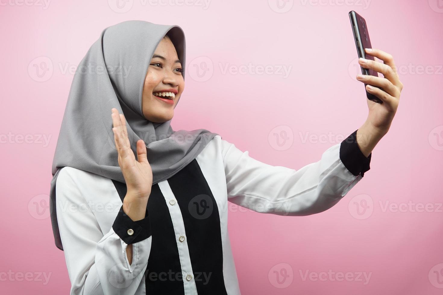 Schöne junge asiatische muslimische Frau, die selbstbewusst, enthusiastisch und fröhlich lächelt, mit den Händen, die Smartphone halten, jemanden anrufen, Videoanrufe tätigen, Selfie machen, einzeln auf rosa Hintergrund foto