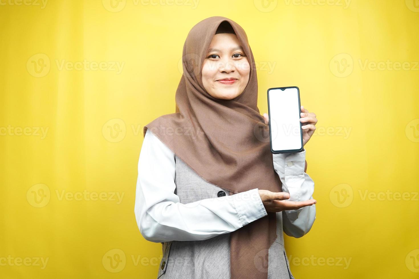 Schöne junge asiatische muslimische Frau, die selbstbewusst und aufgeregt mit den Händen lächelt, die Smartphone halten, App bewerben, etwas fördern, isoliert auf gelbem Hintergrund, Werbekonzept foto