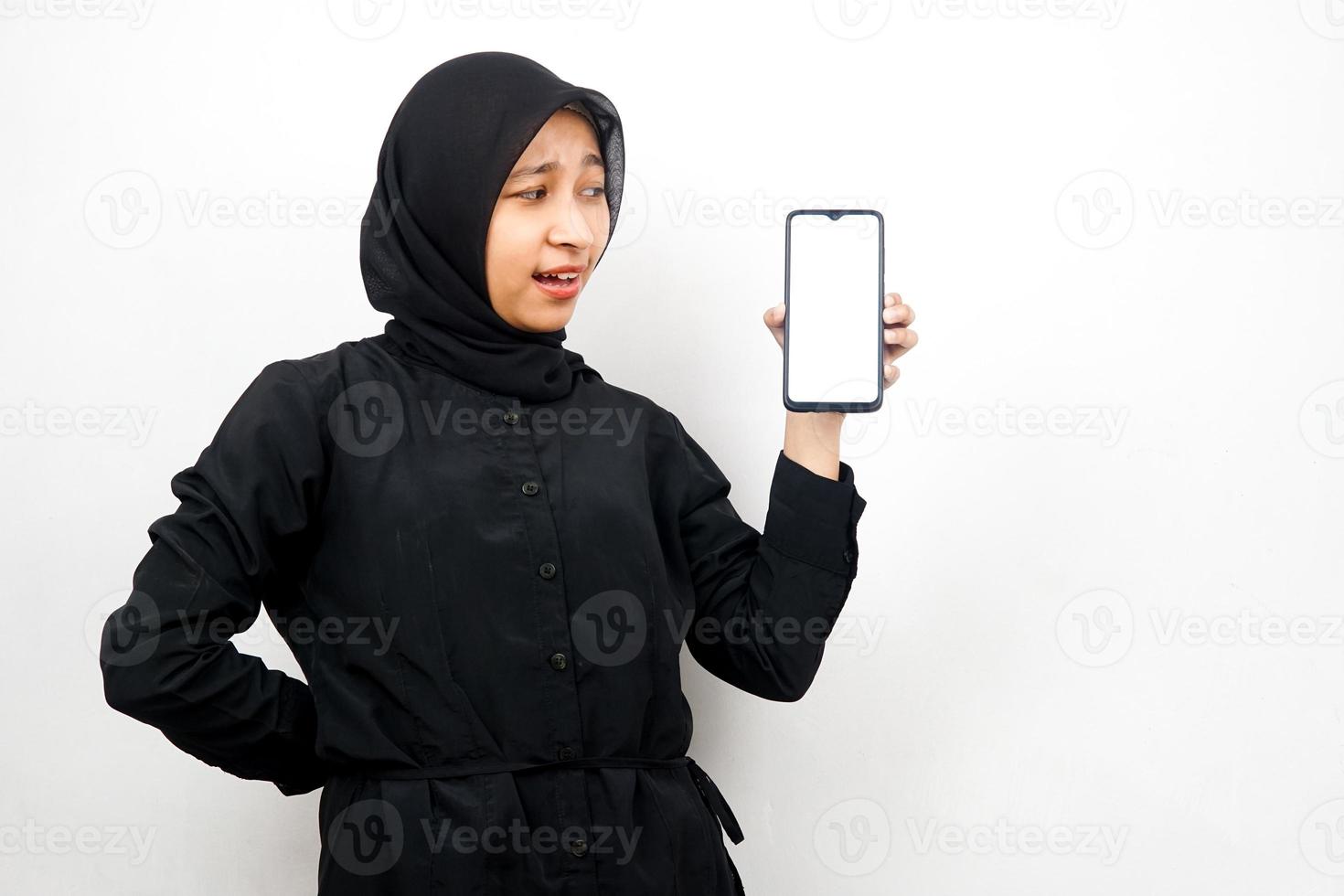 schöne junge asiatische muslimische frau schockiert, überrascht, wow-ausdruck, hand hält smartphone mit weißem oder leerem bildschirm, fördert app, fördert produkt, präsentiert etwas, isoliert foto