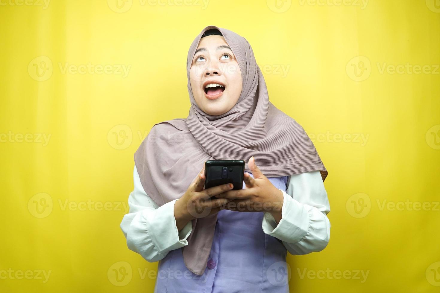 schöne junge asiatische muslimische frau schockiert, überrascht, wow-ausdruck, hand, die smartphone hält, nach oben schauend, isoliert auf gelbem hintergrund foto