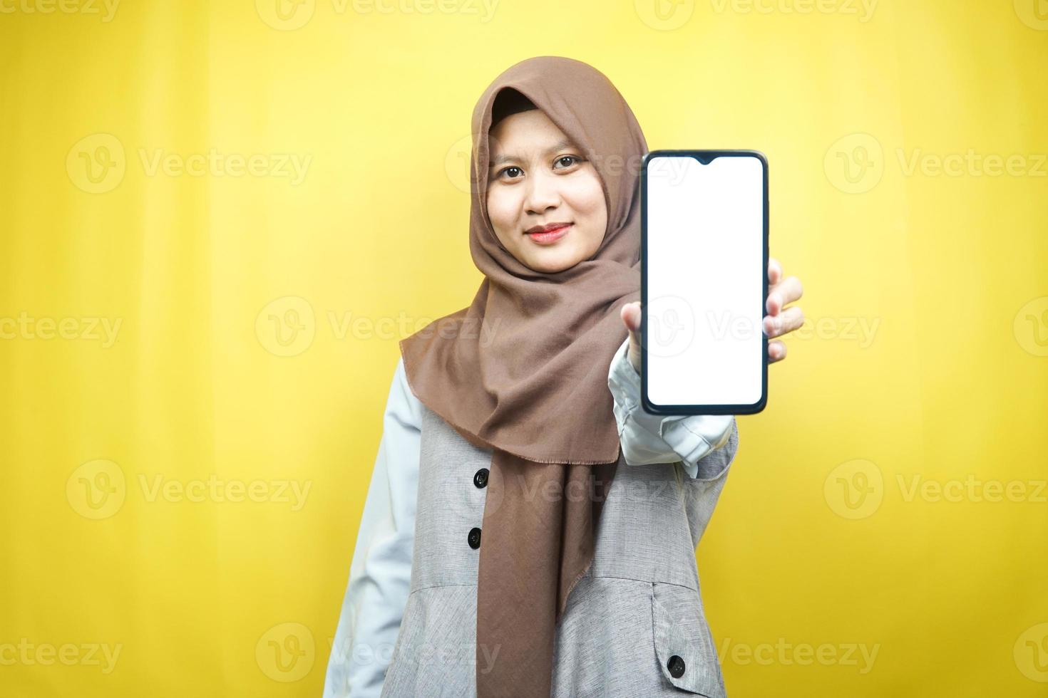 Schöne junge asiatische muslimische Frau, die selbstbewusst, enthusiastisch und fröhlich lächelt, mit der Hand, die das Smartphone hält, etwas fördert, die Hand, die App fördert, einzeln auf gelbem Hintergrund foto