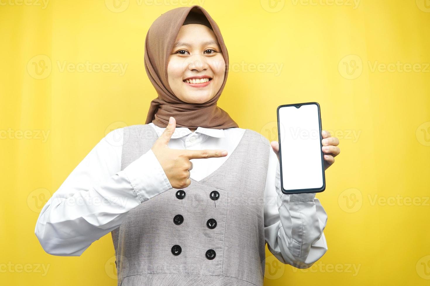 schöne junge asiatische muslimische frau, die selbstbewusst, enthusiastisch und fröhlich lächelt, mit der hand, die auf das smartphone zeigt, etwas fördert, die hand fördert die app, einzeln auf gelbem hintergrund foto