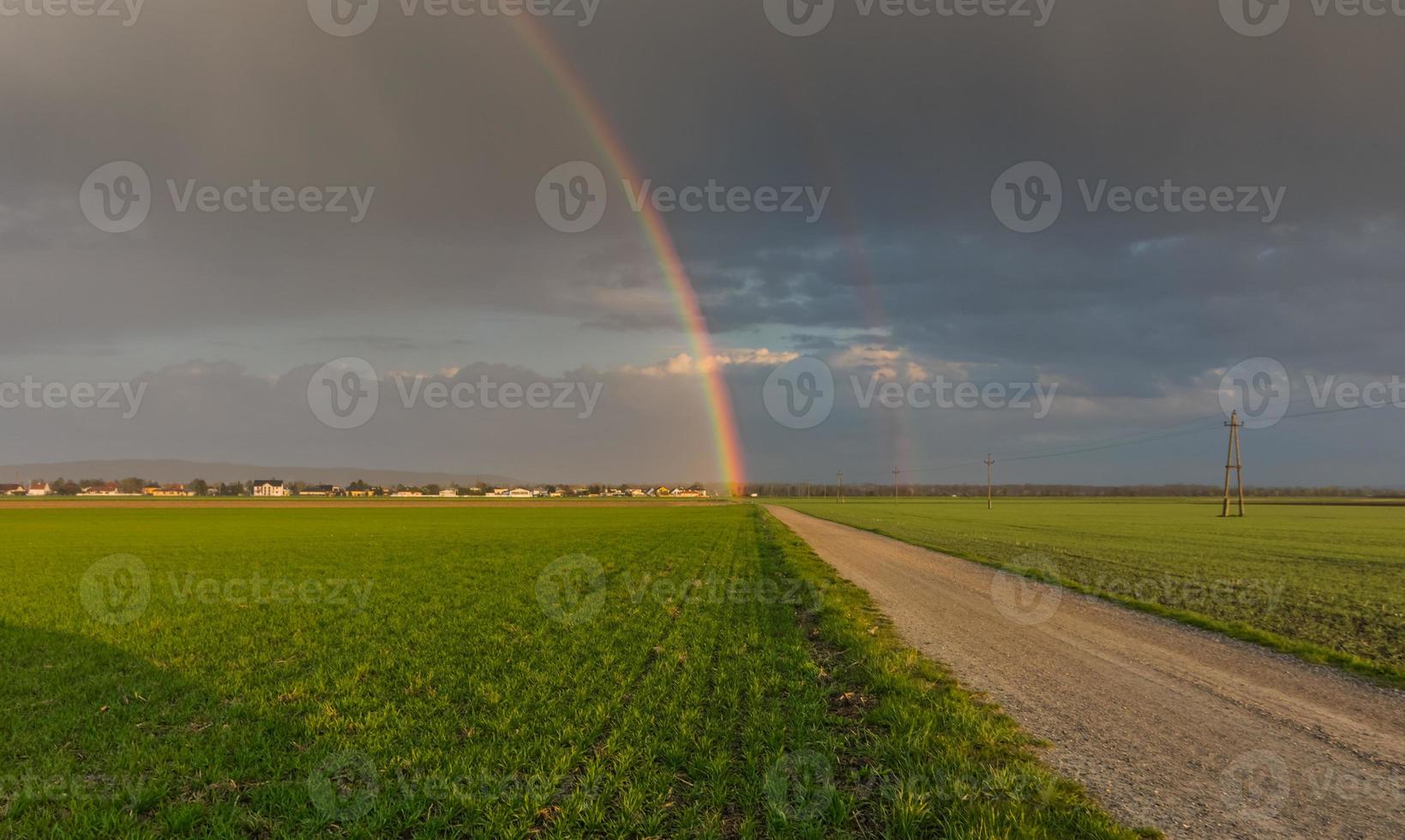 wunderbarer Regenbogen am Ende einer Schotterstraße mit grünen Feldern foto