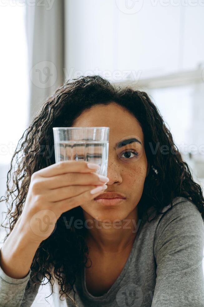 junge schwarze Frau, die Wasserglas betrachtet, während sie in der Küche sitzt foto