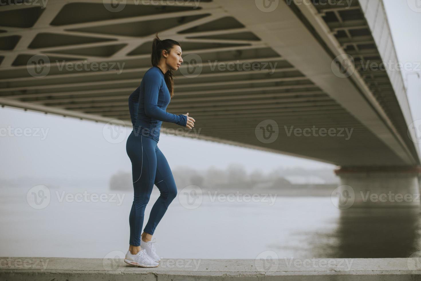 junge Frau im blauen Trainingsanzug, die sich am Herbstmorgen vor dem Training am Fluss ausdehnt foto