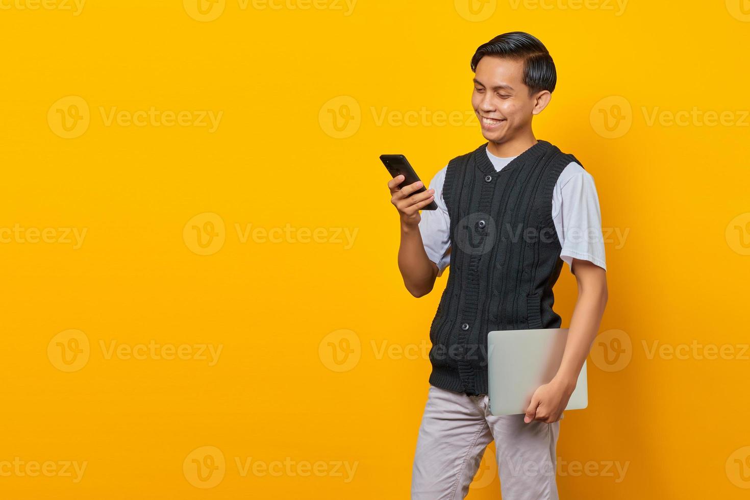 Porträt eines glücklichen jungen asiatischen Mannes, der Laptop hält und Handy auf gelbem Hintergrund benutzt foto