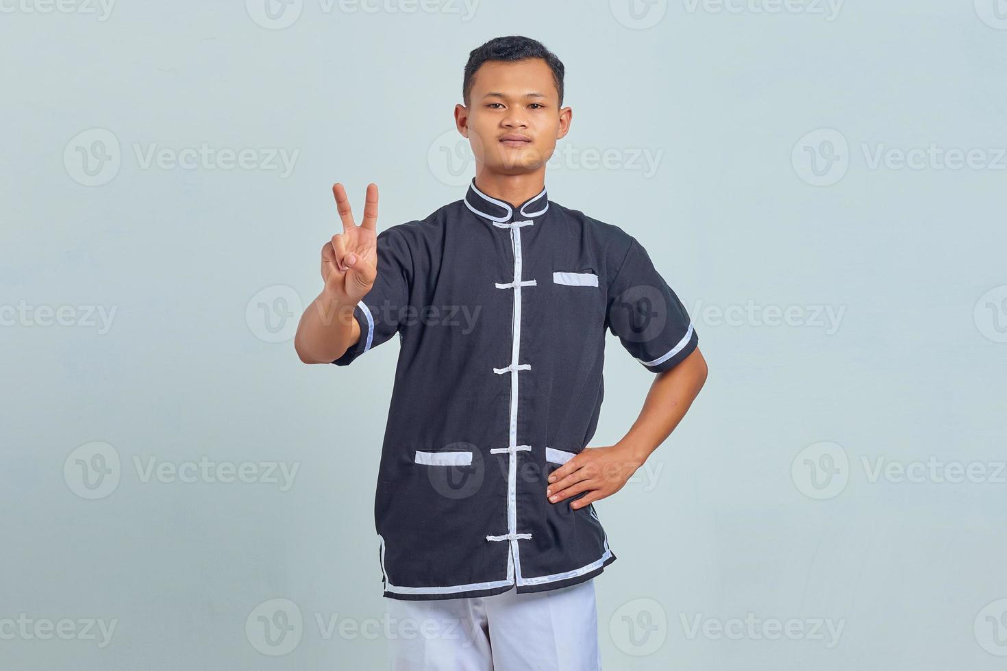 Porträt eines jungen asiatischen Mannes mit Taekwondo-Kimono, der ein Friedenszeichen mit dem Finger auf grauem Hintergrund macht foto