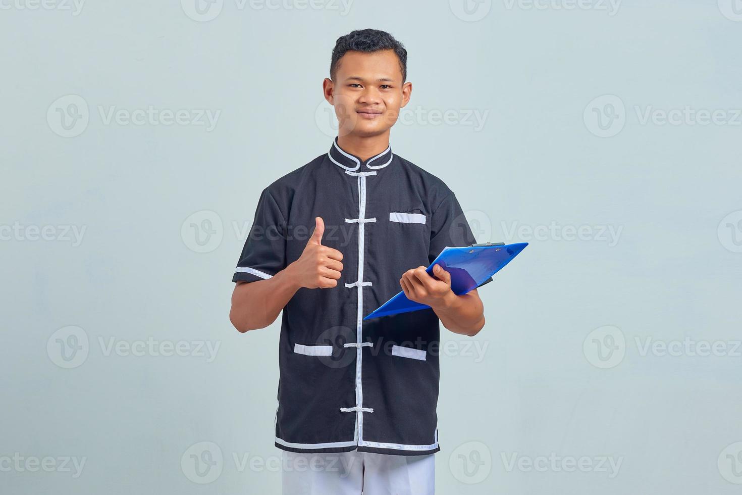 Porträt eines lächelnden asiatischen jungen Mannes in Karate-Uniform, der eine Zwischenablage mit Daumen nach oben auf grauem Hintergrund hält foto