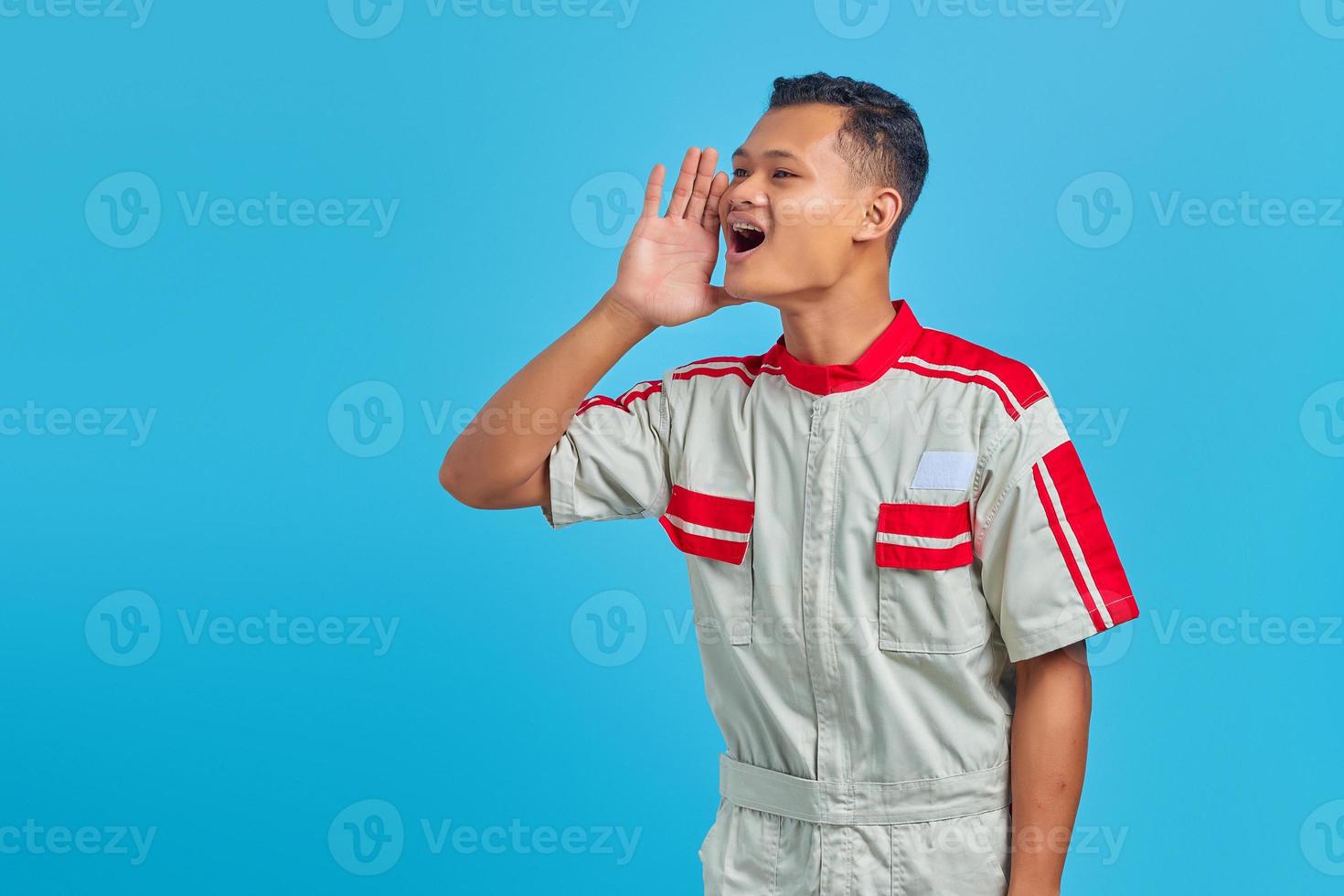 Porträt eines lächelnden jungen asiatischen Mechanikers, der mit der Hand auf dem Mund auf blauem Hintergrund laut seitwärts schreit und schreit foto