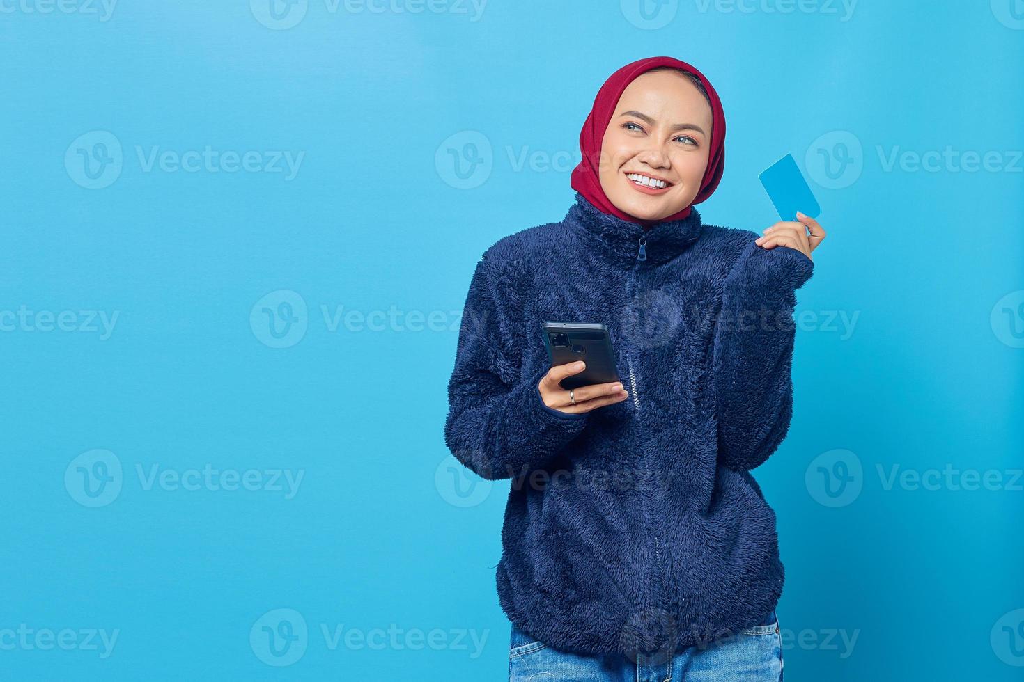 fröhliche junge asiatische frau, die aufschaut, während sie das handy hält und kreditkarte auf blauem hintergrund zeigt foto