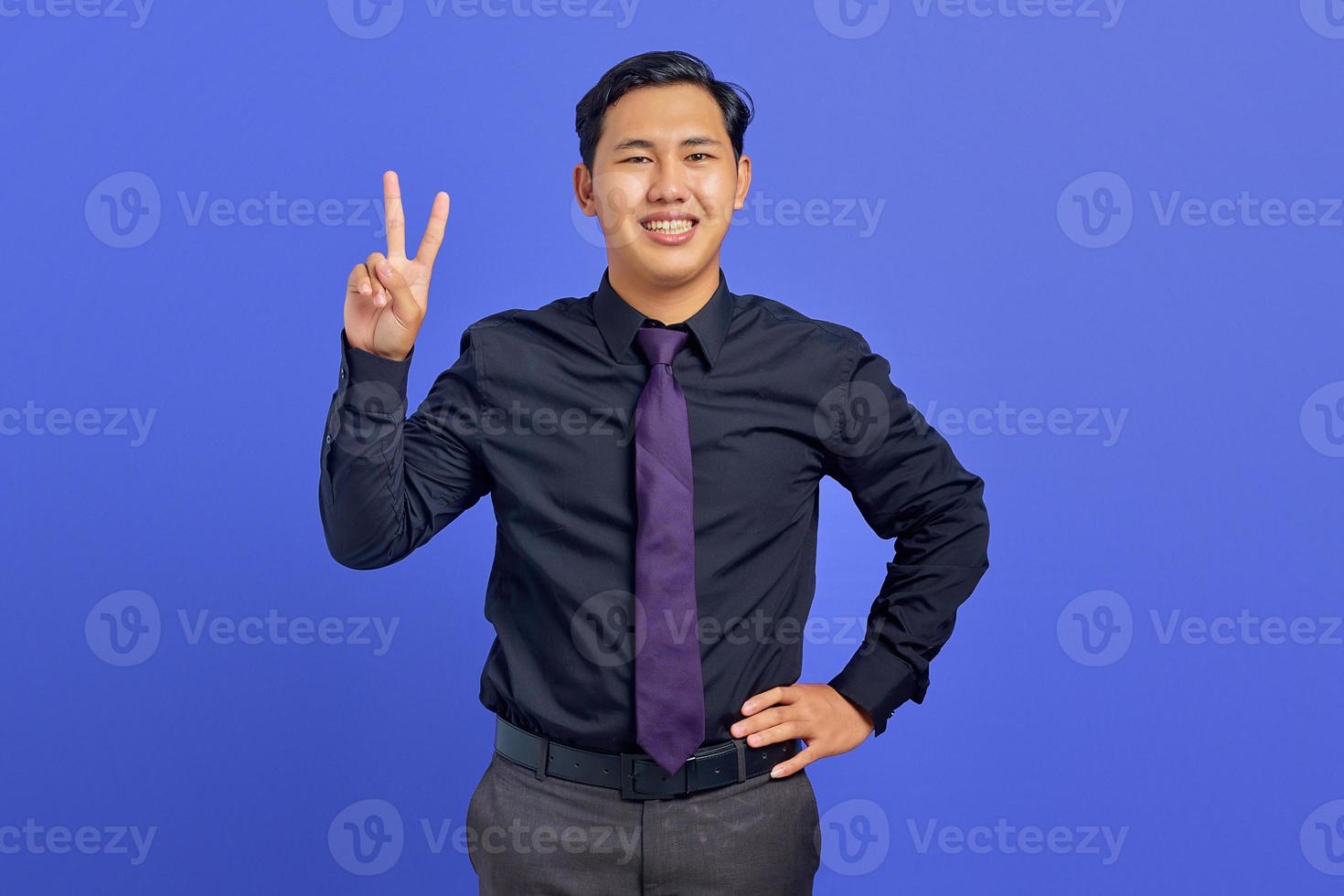 hübscher fröhlicher junger asiatischer mann, der friedenszeichen auf lila hintergrund zeigt foto