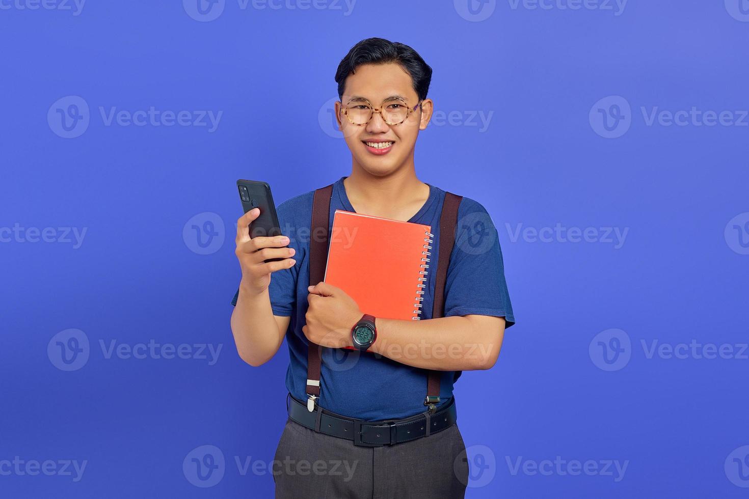 lächelnder junger asiatischer mann, der handy und notizbuch auf violettem hintergrund hält foto