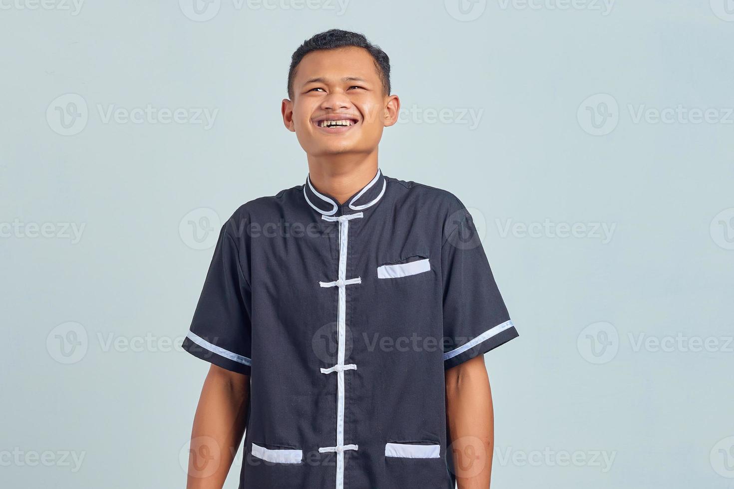 Porträt eines jungen asiatischen Mannes, der Karate-Uniform trägt und über lustigen verrückten Witz lacht, mit den Händen über den Körper auf grauem Hintergrund foto