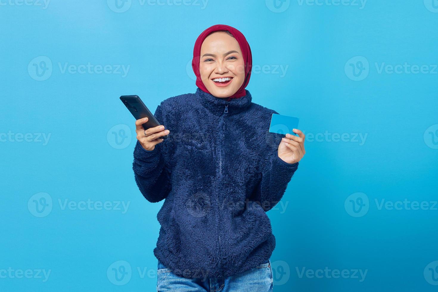Porträt einer lächelnden jungen Asiatin, die Handy hält und Kreditkarte auf blauem Hintergrund zeigt foto