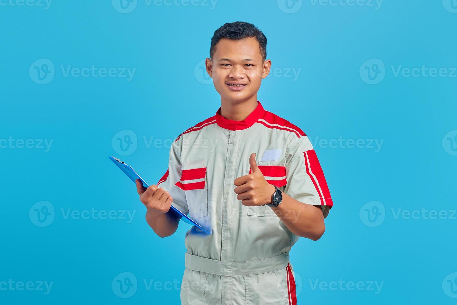 Porträt eines aufgeregten jungen asiatischen Mechanikers, der Zwischenablage hält und Daumen nach oben Geste zeigt, isoliert auf blauem Hintergrund foto