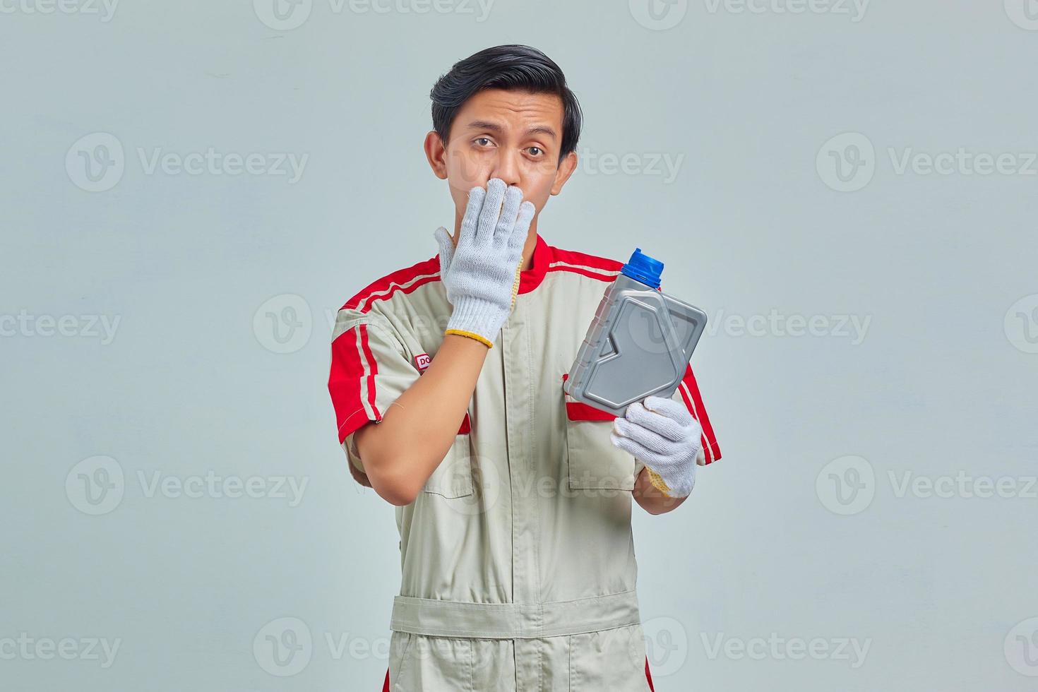 Porträt eines überraschten gutaussehenden Mannes, der eine Mechanikeruniform trägt, die eine Plastikflasche mit Motoröl hält und den Mund mit den Händen auf grauem Hintergrund bedeckt foto