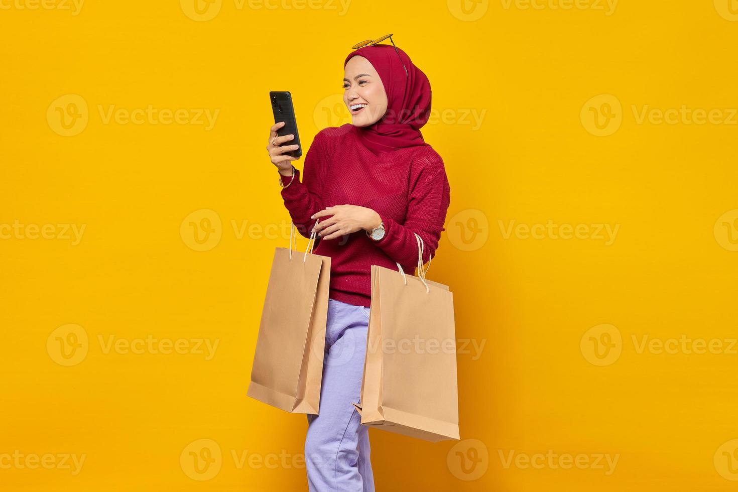 Fröhliche junge Asiatin liest Nachricht auf dem Smartphone und zeigt Einkaufstüten auf gelbem Hintergrund foto