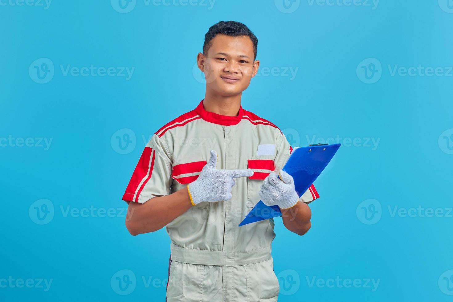 Porträt eines lächelnden jungen asiatischen Mechanikers, der mit dem Finger auf blauem Hintergrund auf die Zwischenablage zeigt foto